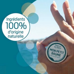 Mon Petit Baume est tenu entre les doigts d'une main réalisant le signe ok. La personne, non visible, est située sur la plage devant les thermes au bord de la lagune de Thau. Une pastille d'information indique sur fond bleu que le petit baume contient 100% d'ingrédients d'origine naturelle